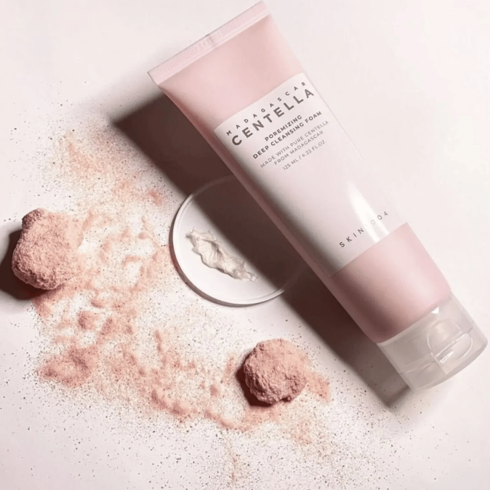 Madagascar Centella Poremizing Deep Cleansing Foam presenteres i en rosa tube. Tuben ligger på et lyst underlag sammen med rosa pulver som gir en følelse av renhet og friskhet. Produktet er designet for å dyprense huden ved hjelp av ren centella fra Madagaskar.
