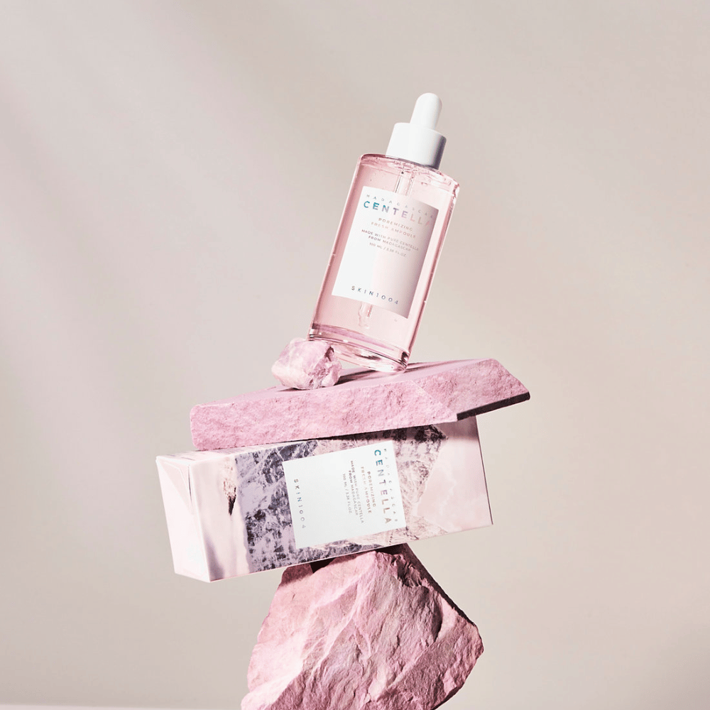 Gjennomsiktig flaske med rosa Centella-ampull fra SKIN1004, balansert på steinblokker, med en kartongforpakning under, mot en nøytral bakgrunn.
