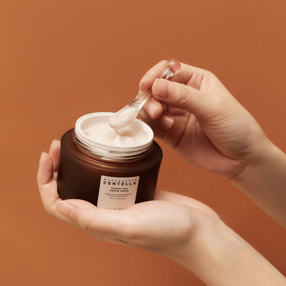 Hender som holder en åpen krukke av Madagascar Centella Probio-Cica Enrich Cream og en spatel med krem mot en enhetlig terrakottafarget bakgrunn.