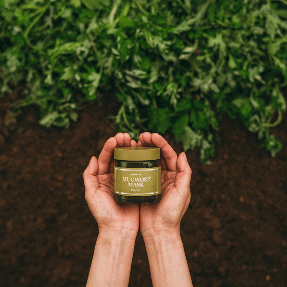 Hender som holder i I'm From Mugwort Mask mot en bakgrunn av jord og friske grønne urter, som fremhever produktets naturlige ingredienser.