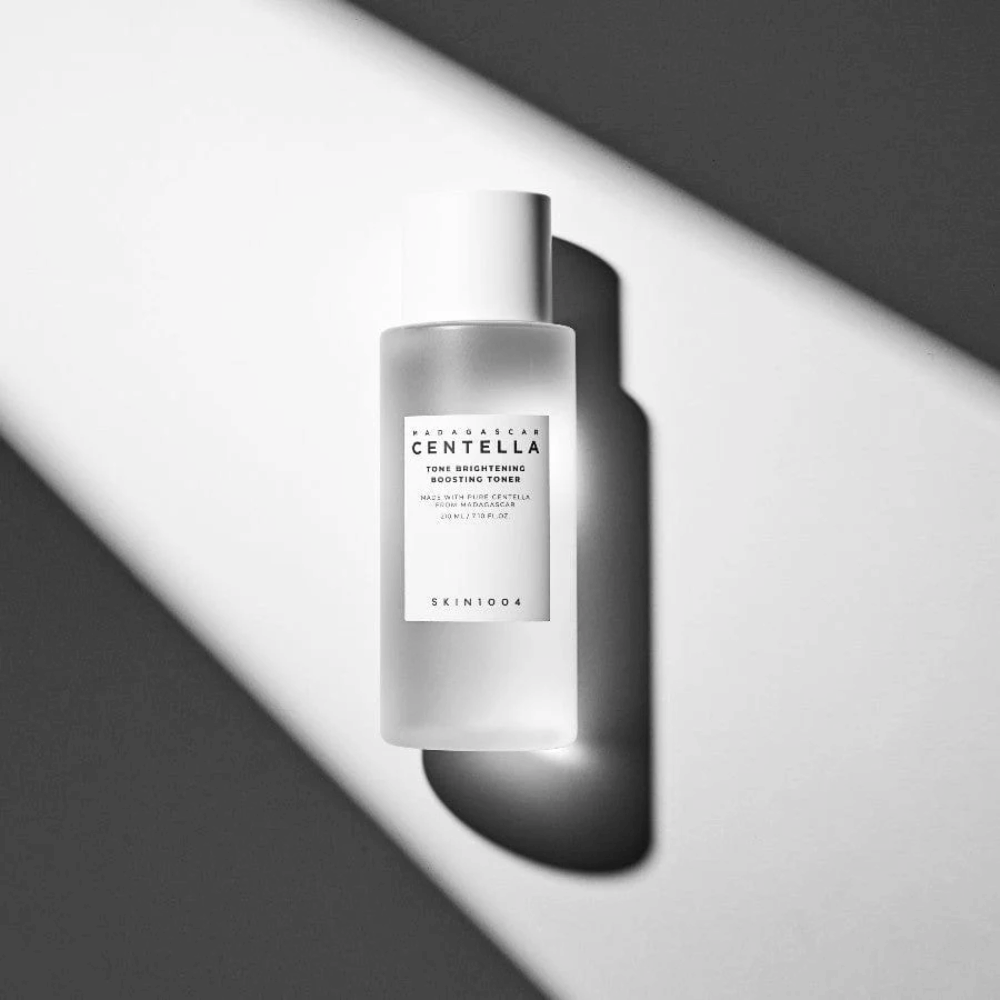 Flaske av SKIN1004 Madagascar Centella Tone Brightening Boosting Toner liggende på hvit overflate med dramatisk skygge i svart-hvitt lys.