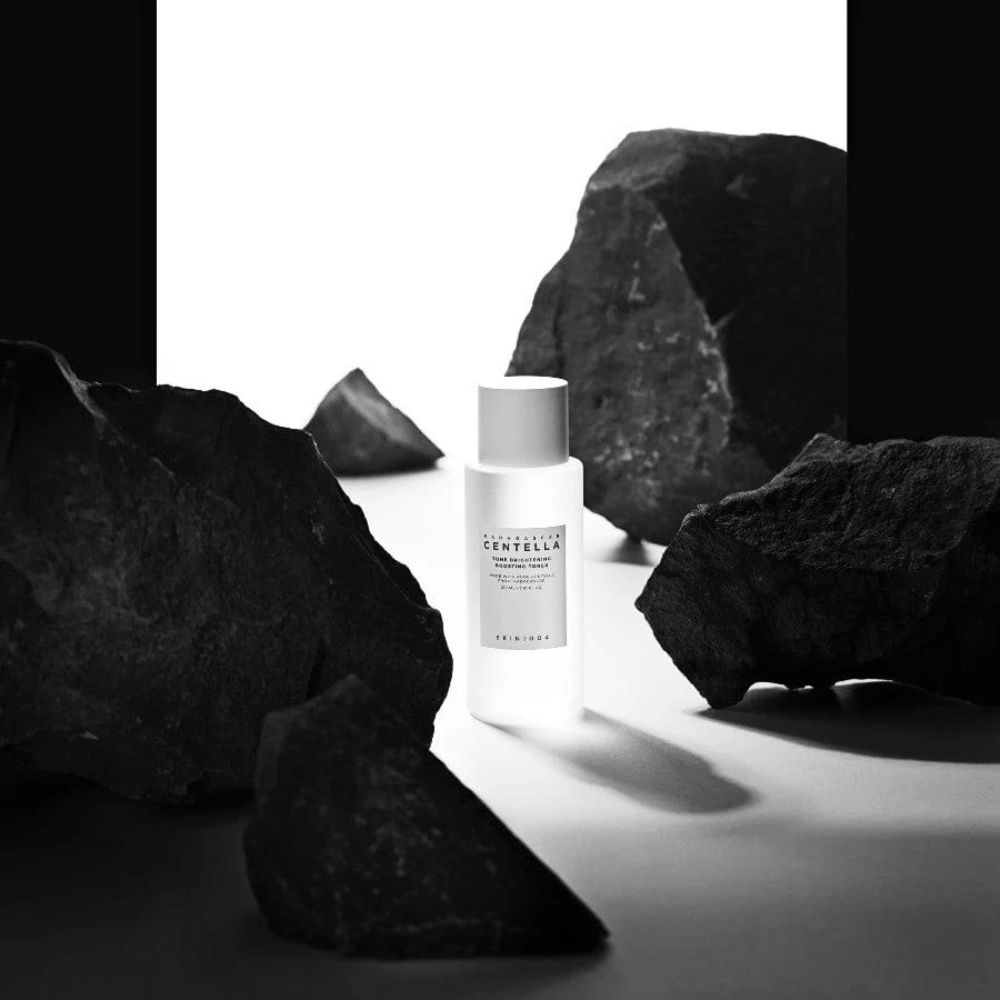 Hvit flaske SKIN1004 Madagascar Centella Tone Brightening Boosting Toner plassert dramatisk blant store svarte steiner mot hvit bakgrunn.
