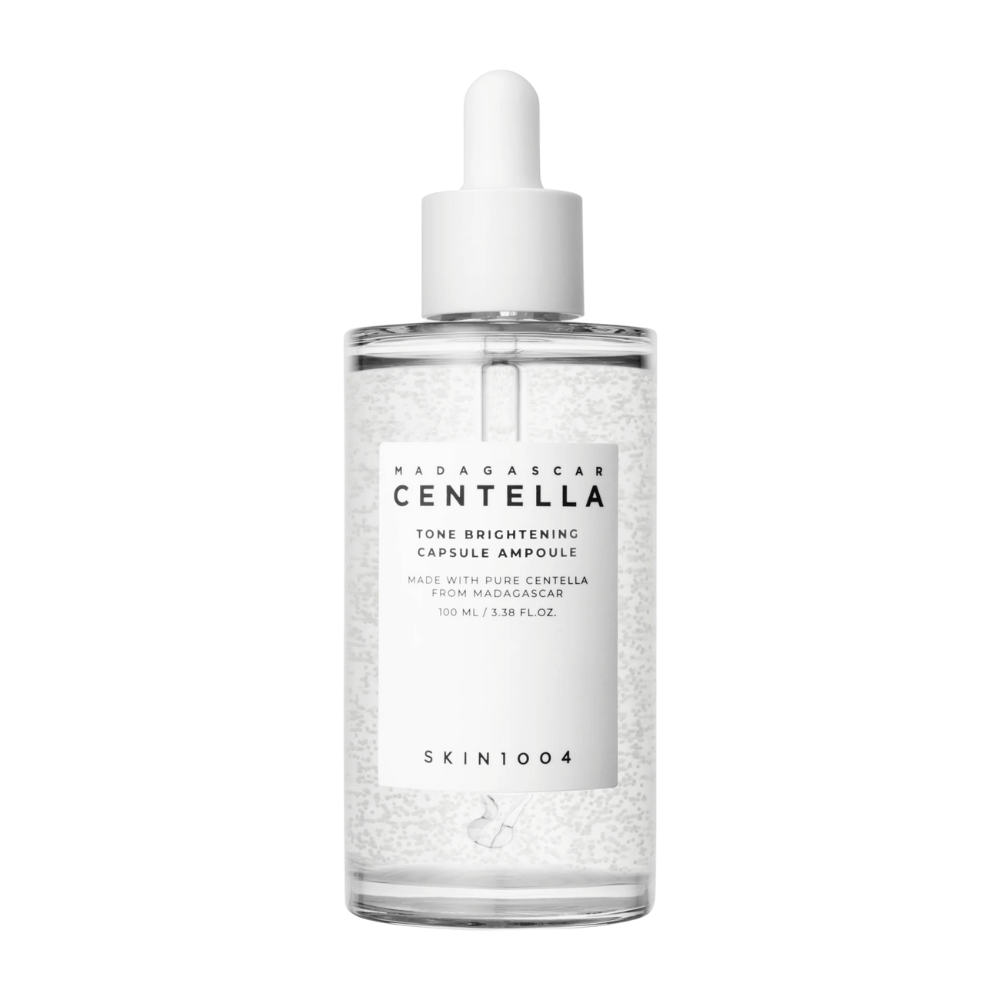 Bildet viser en flaske av "SKIN1004 Madagascar Centella Tone Brightening Capsule Ampoule". Det er en gjennomsiktig flaske med en hvit dråpeapplikator. På etiketten står det at produktet inneholder rene centella-ingredienser fra Madagaskar. Flaskens innhold ser ut til å ha små kapsler eller partikler i seg, noe som kan være en del av dens lysnende hudpleieegenskaper. Flasken inneholder 100 ml / 3.38 fl.oz. av produktet.