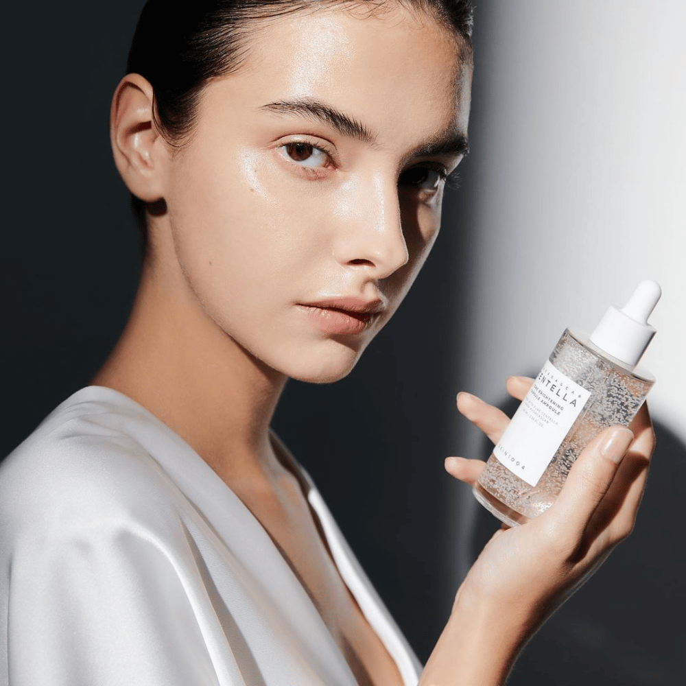 På bildet sees en person som holder en flaske med dråpeapplikator. Flasken er merket "SKIN1004 Madagascar Centella Tone Brightening Capsule Ampoule". Personen har en skimrende hudtone som reflekterer lyset, noe som antyder en mulig effekt av produktet.