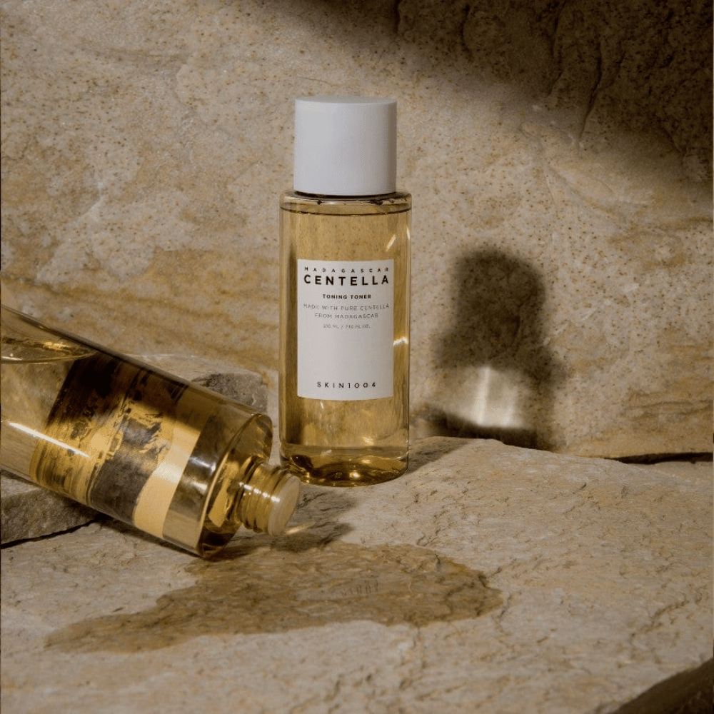 Bildet viser en flaske "Madagascar Centella Toning Toner" fra SKIN1004. Det er en gjennomsiktig flaske fylt med en gulaktig væske, antagelig en toner for hudpleie. Det står at den er laget med ren centella fra Madagaskar og innholdet er 210 ml / 7.10 fl.oz. Flasken og en liggende flaske er plassert på en steinete overflate, noe som gir en naturlig og minimalistisk følelse.