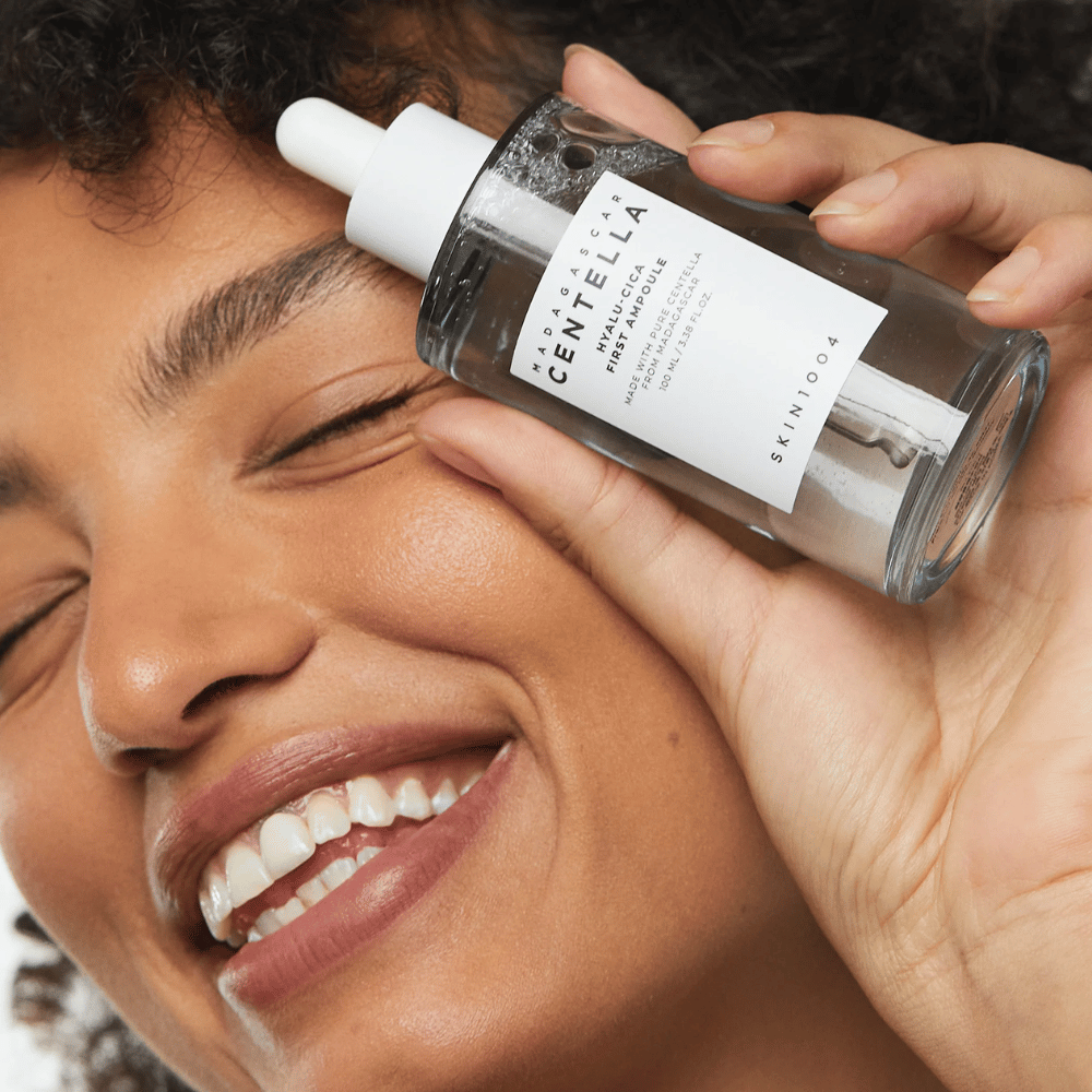 Smilende kvinne holder opp Skin1004 Madagascar Centella Hyalu-Cica First Ampoule ved siden av ansiktet.