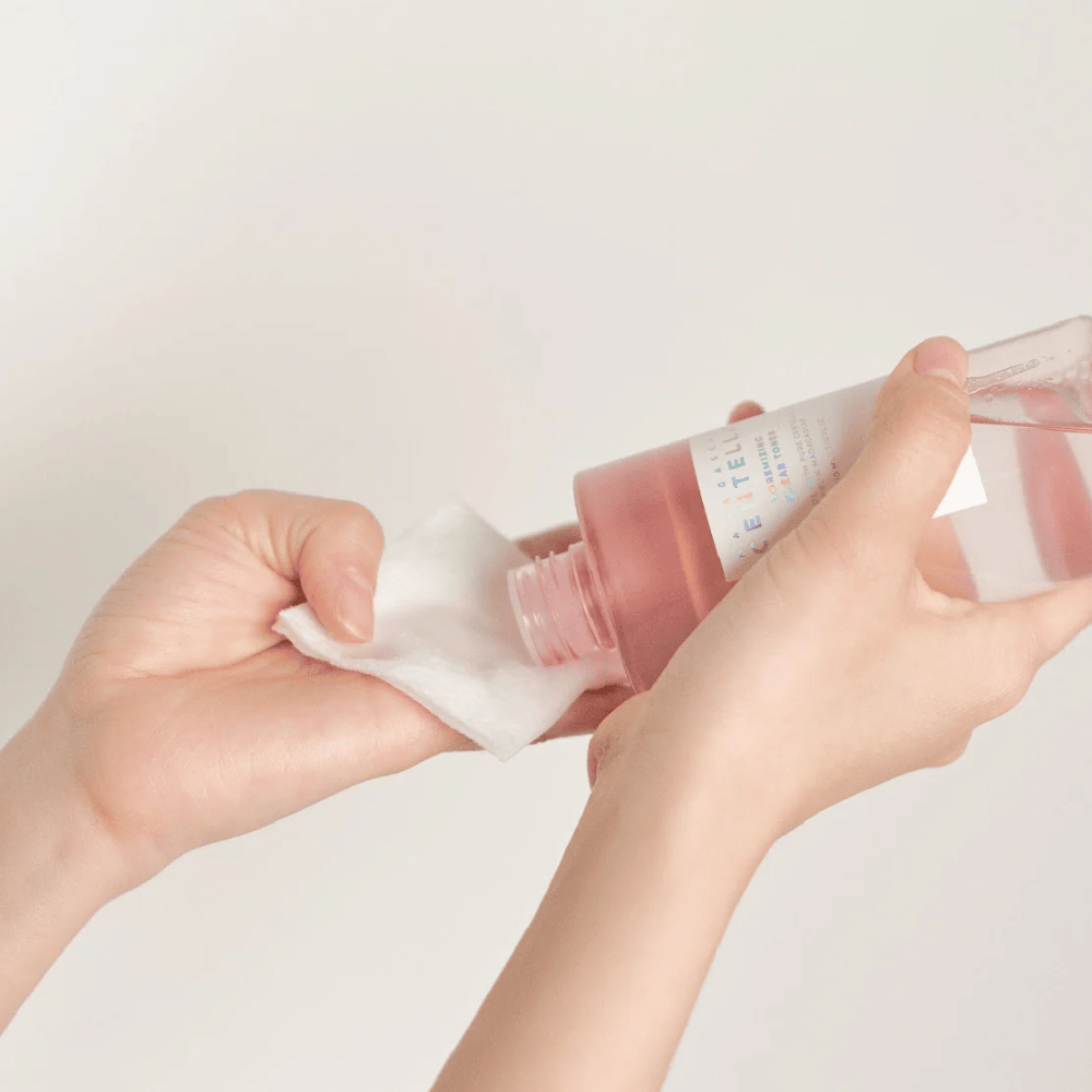 En person heller Madagascar Centella Poremizing Clear Toner fra en transparent flaske på en bomullsrondell. Produktet, som er i en rosa tone, brukes for å friske opp og rense huden. Hendene er synlige, hvilket viser påføringen av toneren.