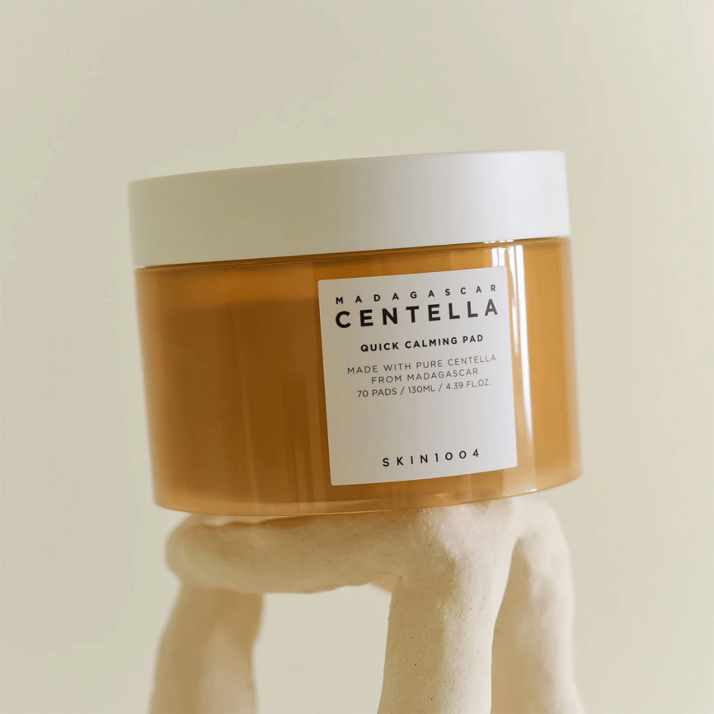 Boks med SKIN1004 Madagascar Centella Quick Calming Pad plassert på en skulpturlignende hvit pidestall – ravfarget beholder med hvit etikett og lokk.