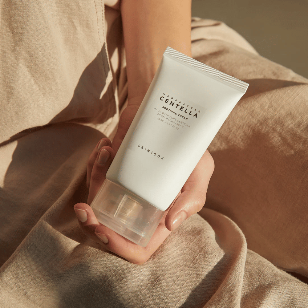 En person holder en hvit tube av Madagascar Centella Soothing Cream fra SKIN1004. Tuben har en gjennomsiktig kork og tekst som beskriver produktet som beroligende og laget med ren Centella. Personen er kledd i lyse klær.