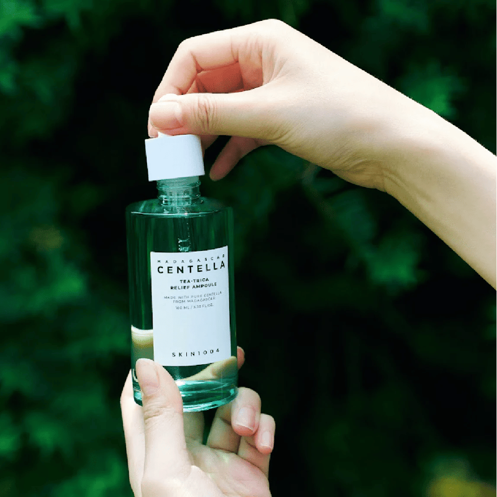 Person åpner SKIN1004 Madagascar Centella Tea-Trica Relief Ampoule – grønn glassflaske med hvit etikett holdes i hender foran frodig bakgrunn.