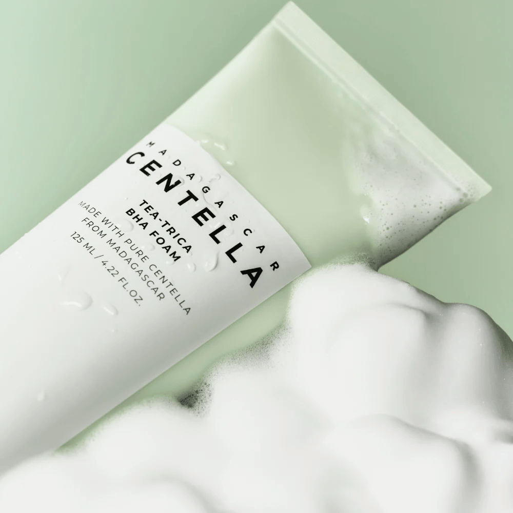 Et produkt av typen renseskum, "Madagascar Centella Tea-trica BHA Foam," er delvis synlig med skum rundt omkring. Produktet har en grønn og hvit design, og teksten "Madagascar Centella" og "Tea-trica BHA Foam" er tydelig synlige. Volumet er 125 ml.