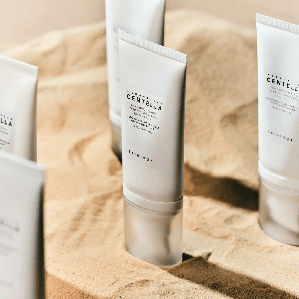 Flere tuber med Madagascar Centella Tone Brightening Tone-Up Sunscreen står i sanden. Tuben er hvit med svart tekst og inneholder 50 ml. Produktet har SPF 50+ PA++++ og er laget med ren Centella fra Madagaskar.