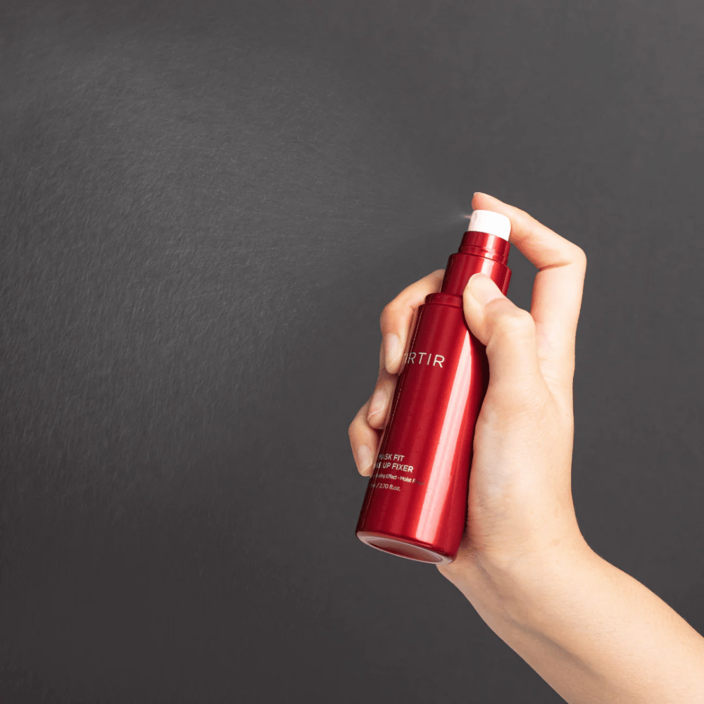 En hånd som holder en rød flaske av merket tirtir Mask Fit Make Up Fixer og sprayer en fin mist, ideell for å fikse sminke.