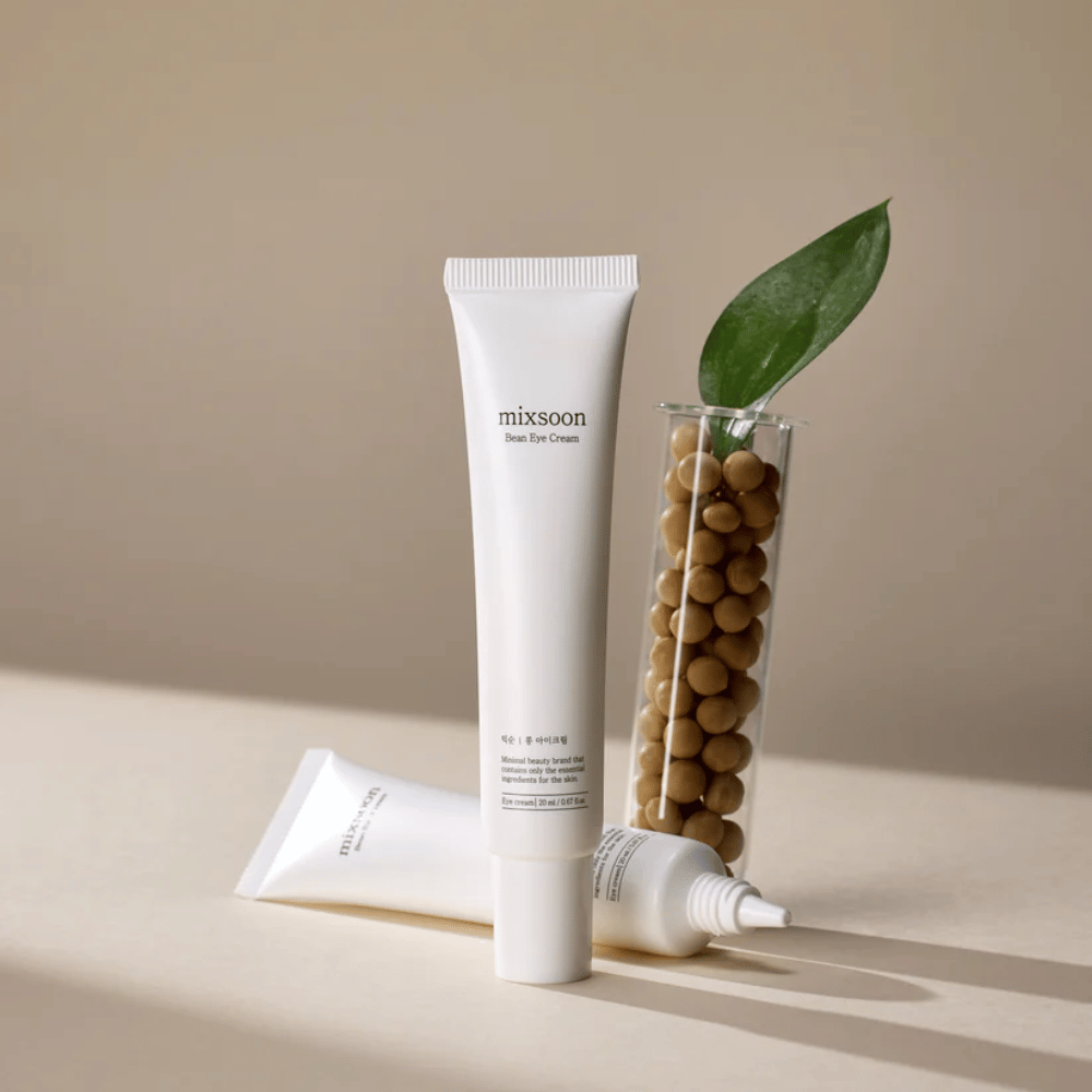 To tuber Mixsoon Bean Eye Cream plassert ved siden av et prøverør fylt med soyabønner og et grønt planteblad, på en beige overflate med skyggespill.