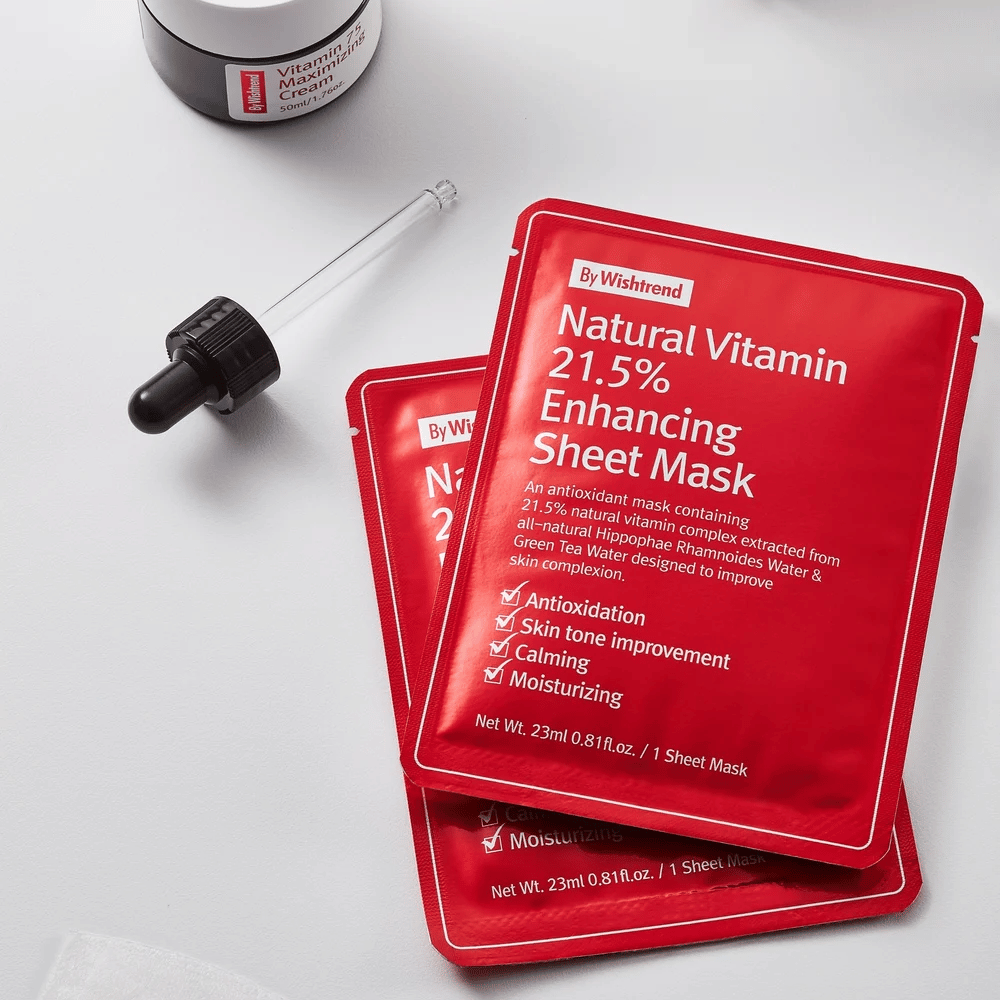 By Wishtrend Natural Vitamin 21.5% Enhancing Sheet mask i røde forpakninger, plassert på en hvit overflate sammen med en pipette og en krukke ansiktskrem.