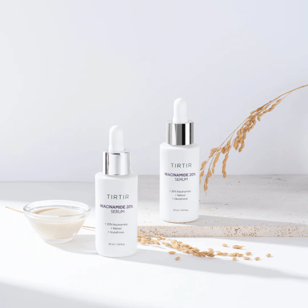 To flasker av Tirtirs "Niacinamide 20% Serum" vises på en lys overflate sammen med en liten skål med en kremaktig substans og noen strå av tørre planter. Serumet inneholder 20 % niacinamid, retinol og glutation, som bidrar til å forbedre hudens utseende og tekstur.