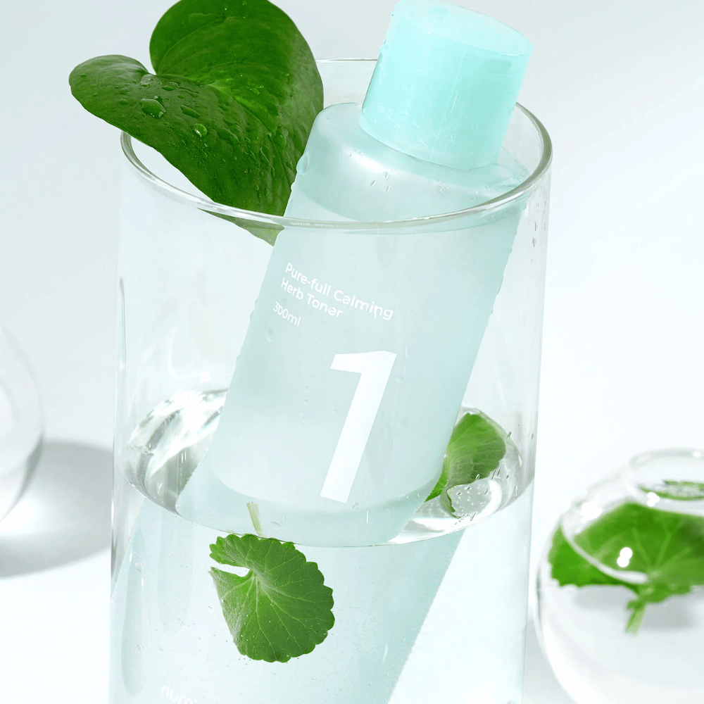 En flaske med No.1 Pure-Full Calming Herb Toner plassert i et glass med vann og grønne blader. Flasken har en lyseblå kork og en etikett med teksten "Pure-full Calming Herb Toner" samt "300ml". Produktet ser friskt ut med vann og vegetasjon i bakgrunnen, noe som gir en beroligende og naturlig følelse.