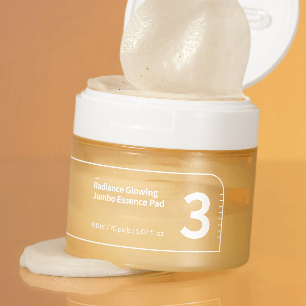 Åpen boks med numbuzin No. 3 Radiance Glowing Jumbo Essence Pad, en pad synlig lent mot kanten, bakgrunn i oransje tone.