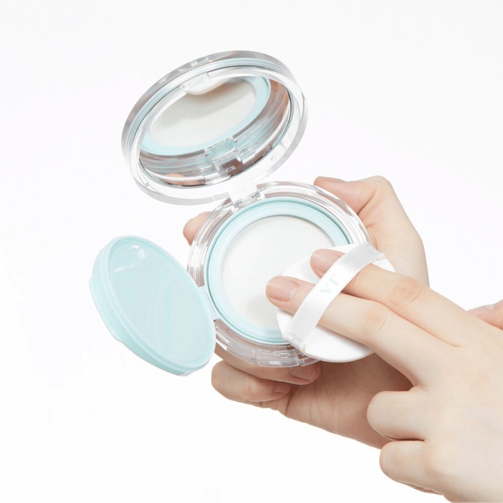 En hånd holder en åpen boks med VT PDRN Essence Glow Sun Pact. Puffen er på vei til å påføre pudderet, som har en hvit overflate og en blå kant rundt boksen.