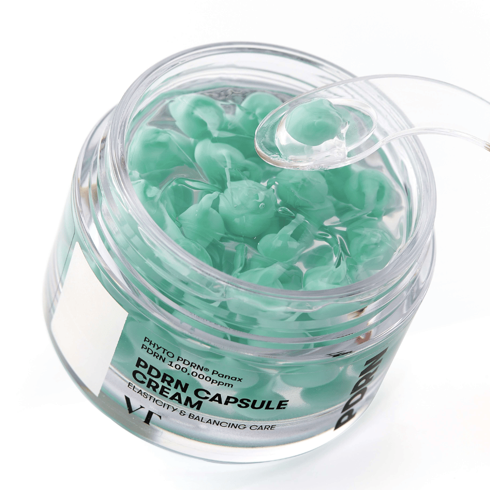 Nærbilde av en åpen krukke med VT PDRN Capsule Cream. Innholdet består av små grønne kapsler i en gjennomsiktig gel, med en medfølgende spatel for påføring. Teksten på krukken lyder "Elasticity & Balancing Care".