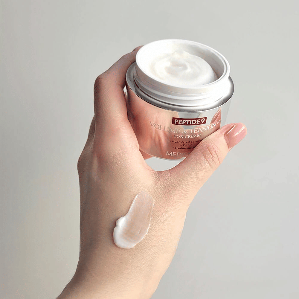 Hånd holder i MEDI-PEEL Peptide 9 Volume & Tension Tox Cream med en klikk krem på håndryggen. Den silkemyke formelen fukter og gir huden fasthet og spenst.