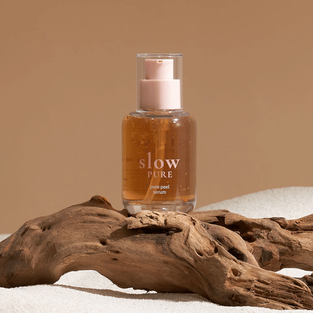 Slow Pure Pore Peel Serum plassert på sand med tredetalj i bakgrunnen, symbol for naturlig hudpleie.