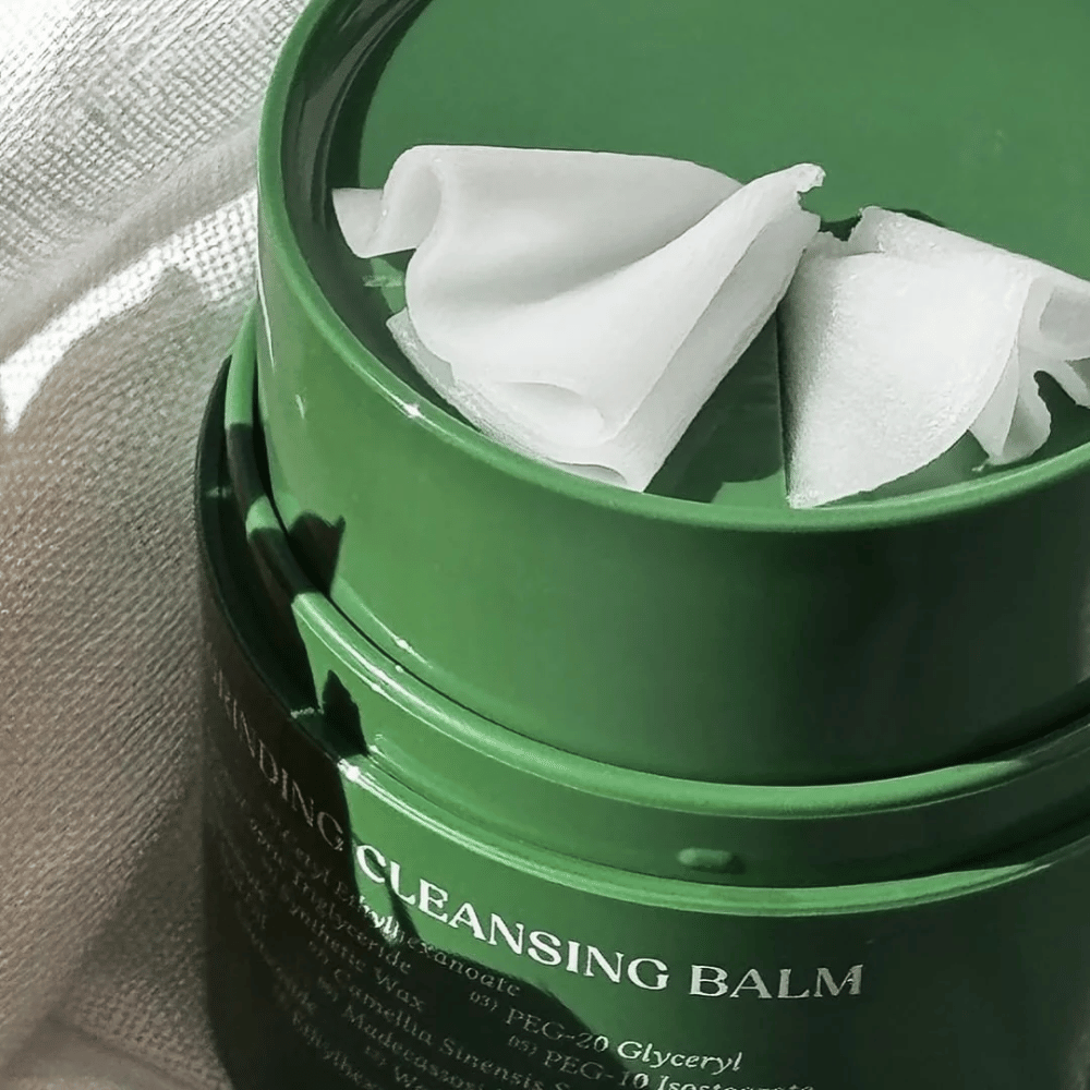 Bildet viser en grønn boks med "Cleansing Balm", som er åpnet for å avsløre den medfølgende spatelen og det myke innholdet. Den kremete konsistensen reflekterer lyset, noe som gir en følelse av luksus og effektivitet i rengjøringen.