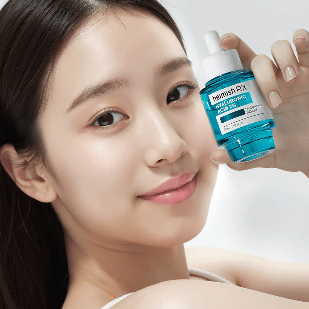 Smilende kvinne med strålende hud holder en flaske Heimish RX Hyaluronic Acid 2% Hydrating Serum, som fremhever dens fuktighetsgivende egenskaper.
