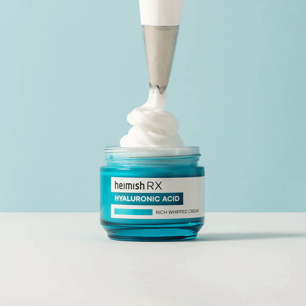 Heimish RX Hyaluronic Acid Rich Whipped Cream i en blå glasskrukke, hvor en sprøytepose påfører den luftige kremen. En fuktighetsgivende ansiktskrem med hyaluronsyre for intensiv fuktighet og en myk, fyldig hud.