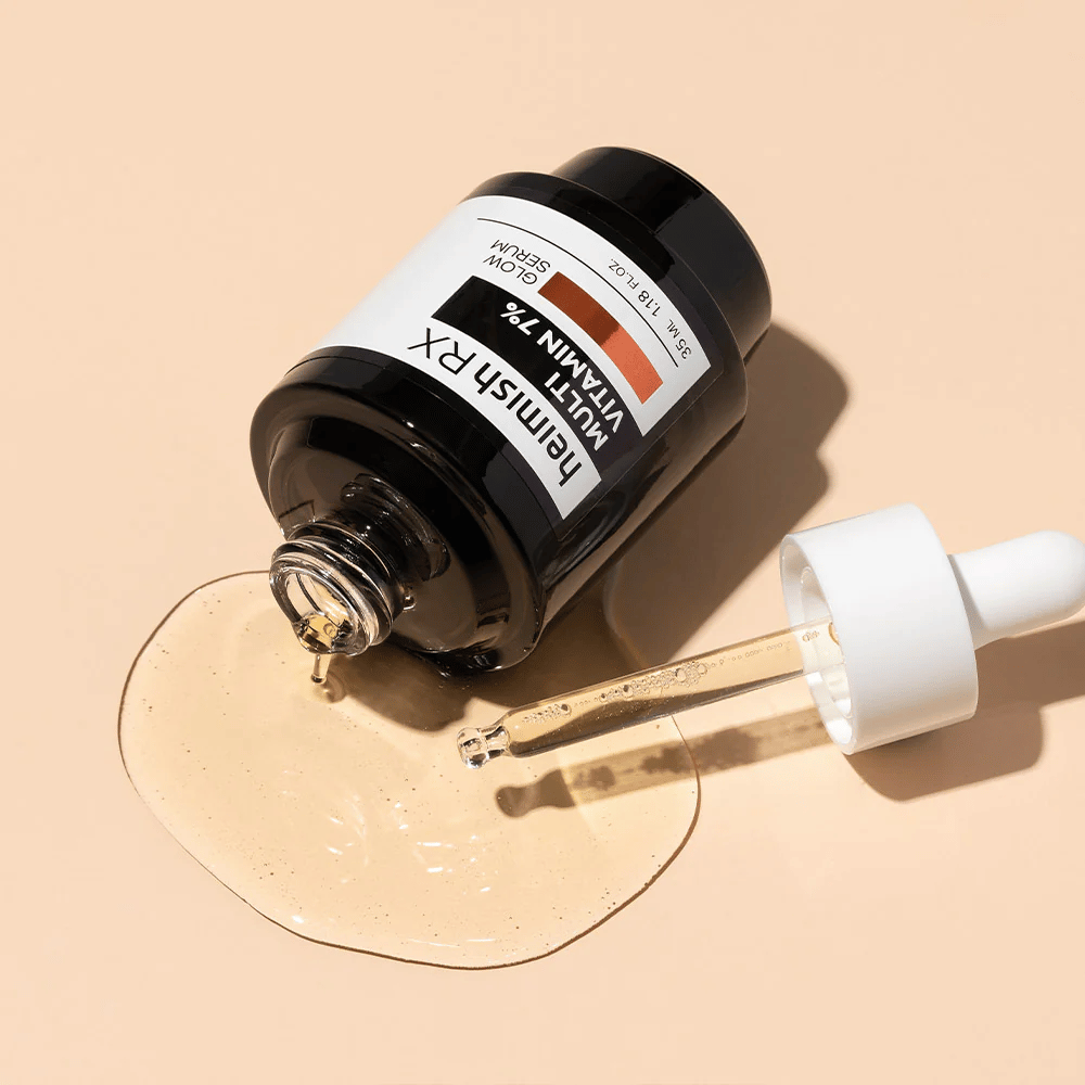 Heimish RX Multi Vitamin 7% Glow Serum liggende på en beige bakgrunn med serum spredt rundt flasken og en pipette fylt med produkt, som fremhever dens næringsrike formel.