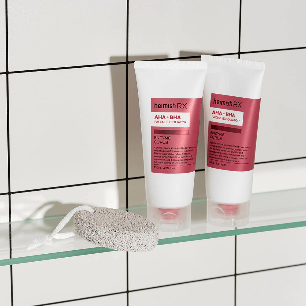 To tuber Heimish RX AHA + BHA Facial Exfoliator plassert på en glasshylle i et bad med hvit flisvegg. Ved siden av produktene ligger en pimpstein med en hvit snor. Et stilrent og minimalistisk hudpleiebilde.