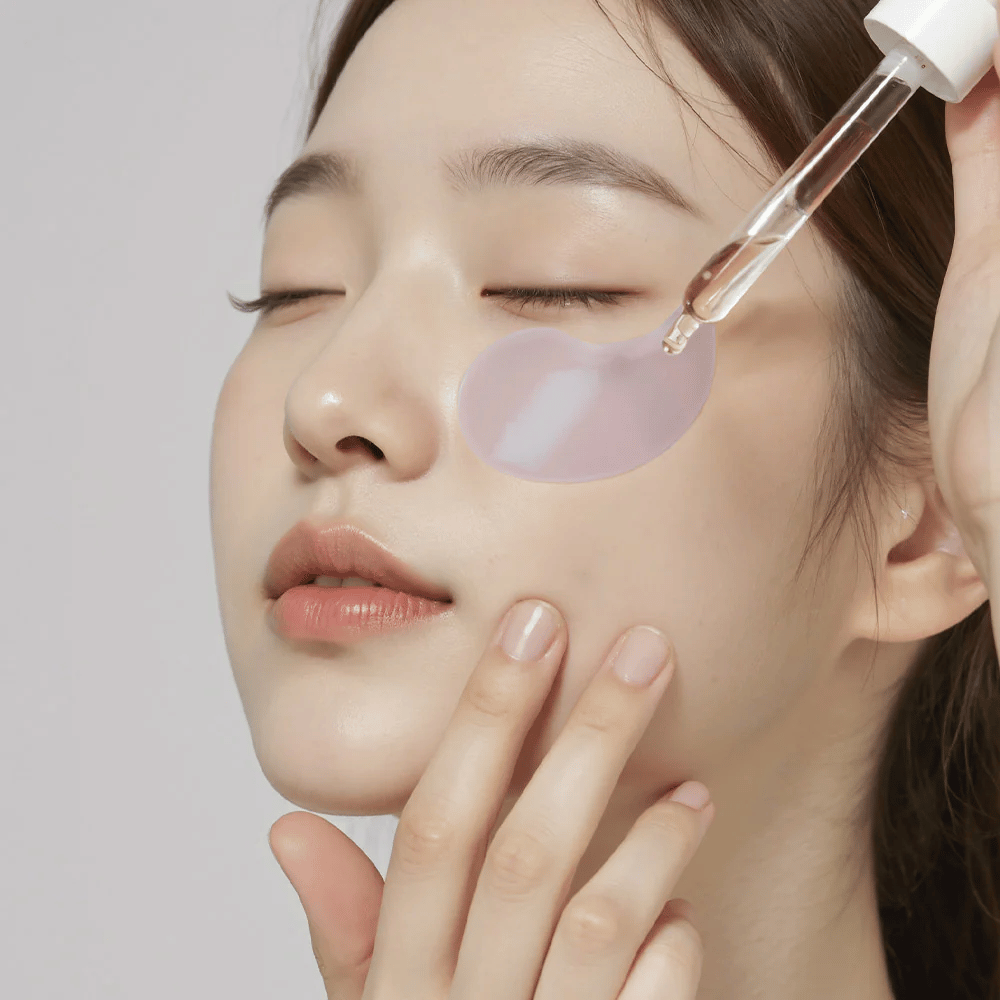 En kvinne med en lilla hydrogel-øyepute påfører serum under øyet med en pipette. Produktet fukter og pleier den sensitive huden rundt øynene.