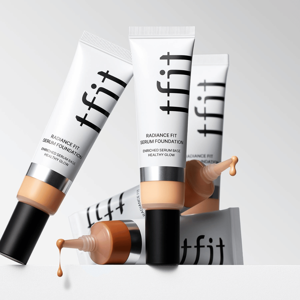 Flere tuber av TFIT Radiance Fit Serum Foundation i forskjellige nyanser, hvorav en med foundation som drypper ut, mot lys grå bakgrunn.