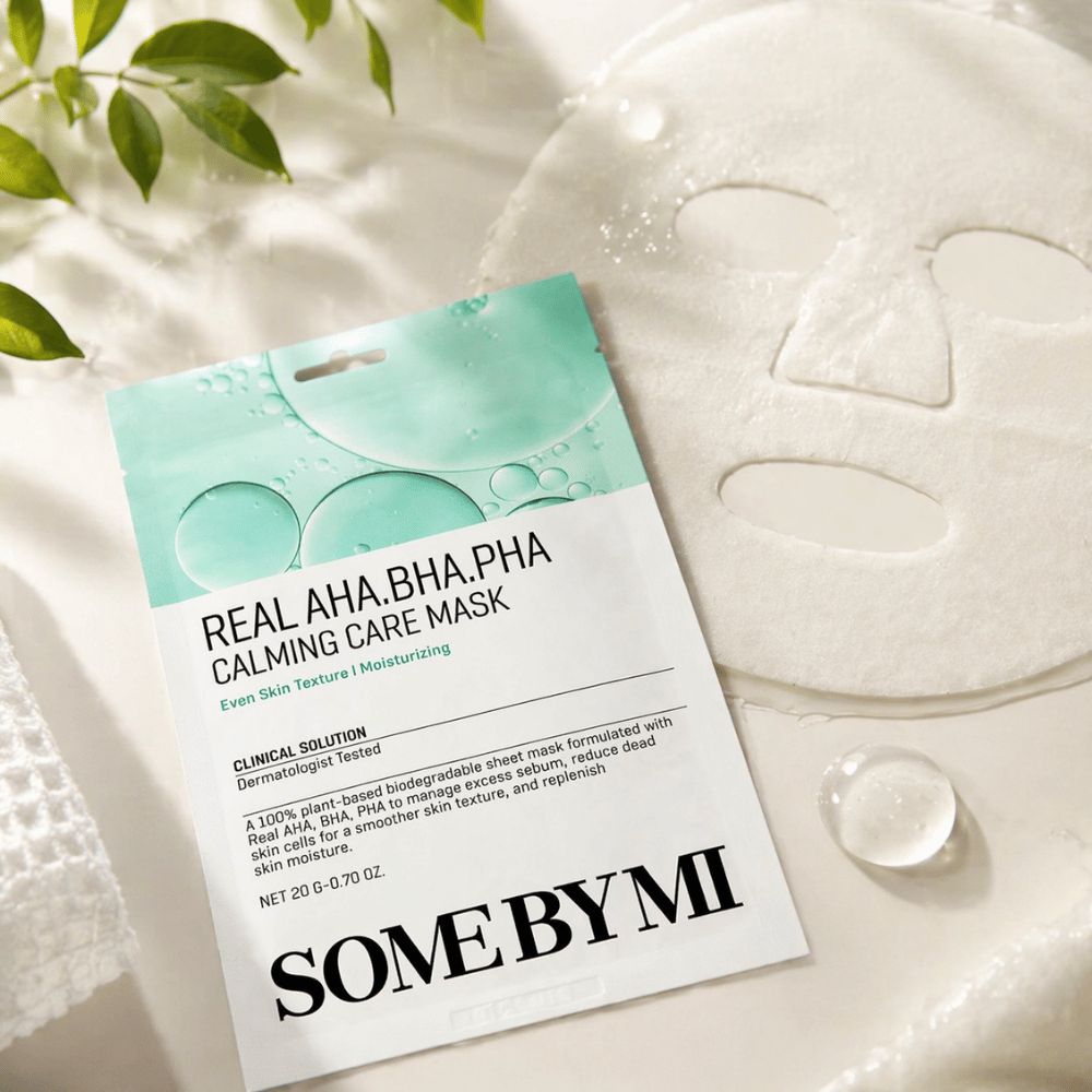 SOME BY MI Real AHA.BHA.PHA Calming Care Mask vises sammen med den utfoldede arkmasken. Rundt produktet sees fuktdråper og grønne blader som forsterker følelsen av fuktighet, beroligende pleie og skånsom eksfoliering. Bildet fremhever maskens fokus på jevnere hudtekstur og forbedret fuktbalanse.
