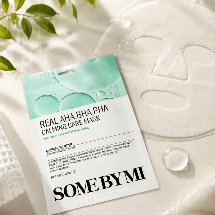 SOME BY MI Real AHA.BHA.PHA Calming Care Mask vises sammen med den utfoldede arkmasken. Rundt produktet sees fuktdråper og grønne blader som forsterker følelsen av fuktighet, beroligende pleie og skånsom eksfoliering. Bildet fremhever maskens fokus på jevnere hudtekstur og forbedret fuktbalanse.