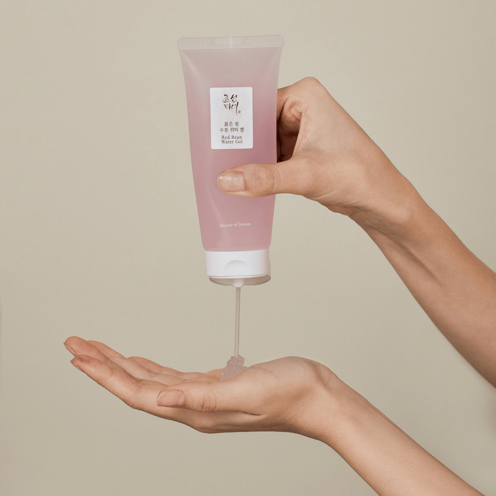 Bildet viser en person som holder et rør av "Red Bean Water Gel" fra Beauty of Joseon og klemmer ut en gjennomsiktig gel i hånden sin. Produktet ser ut til å være beregnet for hudpleie og har en attraktiv, myk rosa emballasje.