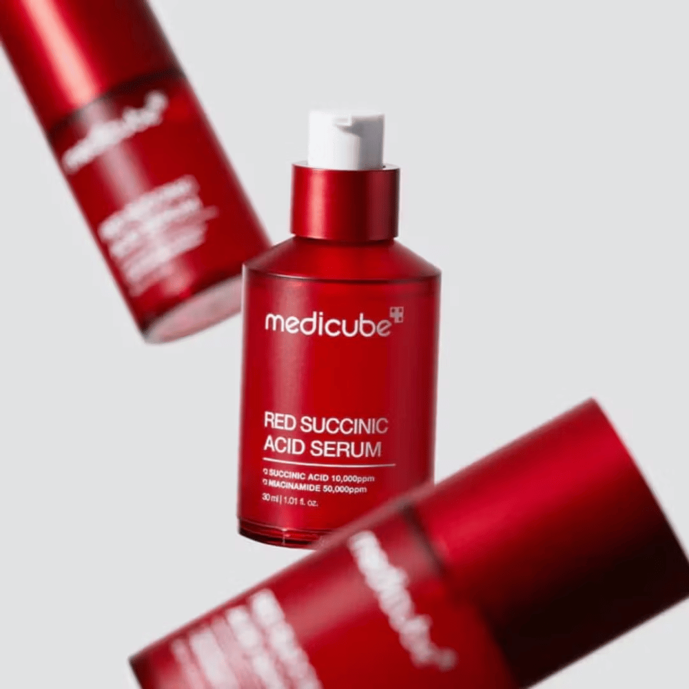 Medicube Red Succinic Acid Serum i rød pumpeflaske, omgitt av flere identiske flasker mot lys bakgrunn. Produktet inneholder succinic acid og niacinamide for å behandle akne og pigmentflekker.