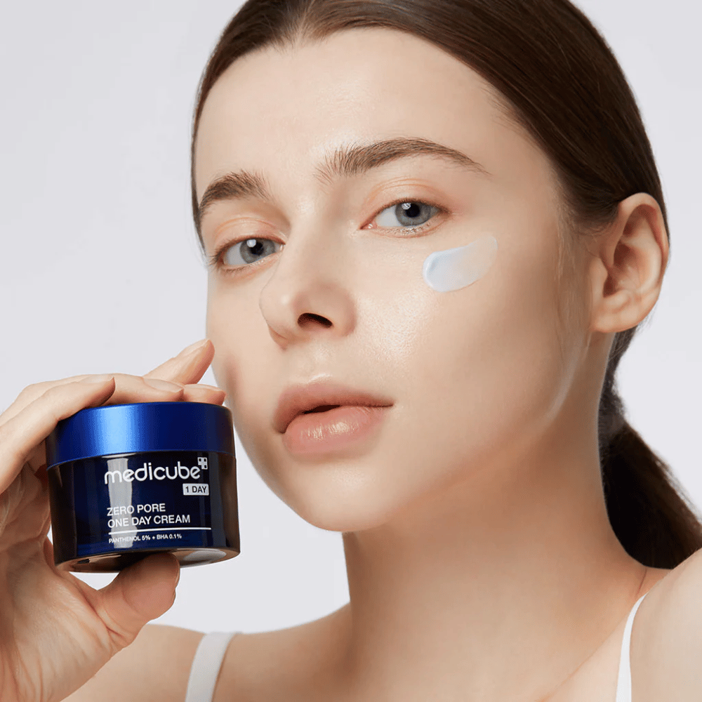 Kvinne med et naturlig utseende holder Medicube Zero Pore One Day Cream. En liten mengde krem er påført hennes kinn. Produktet er designet for å forbedre hudens struktur og minimere porestørrelse, med en lett og fuktighetsgivende formel.