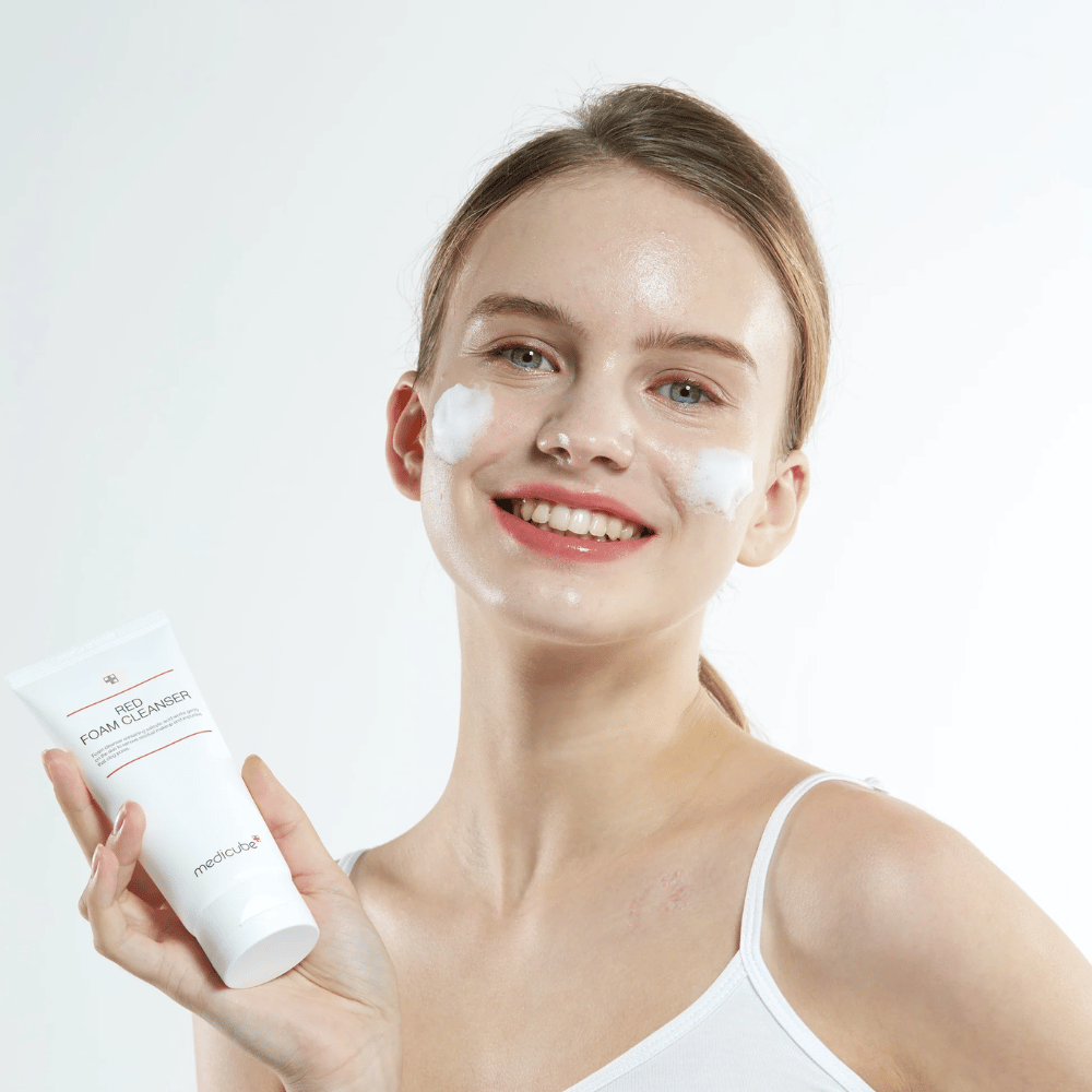 Ung kvinne med rensekum i ansiktet holder opp en tube av Medicube Red Foam Cleanser og smiler mot kameraet. Hun har på seg en hvit singlet og bakgrunnen er lys og enkel.