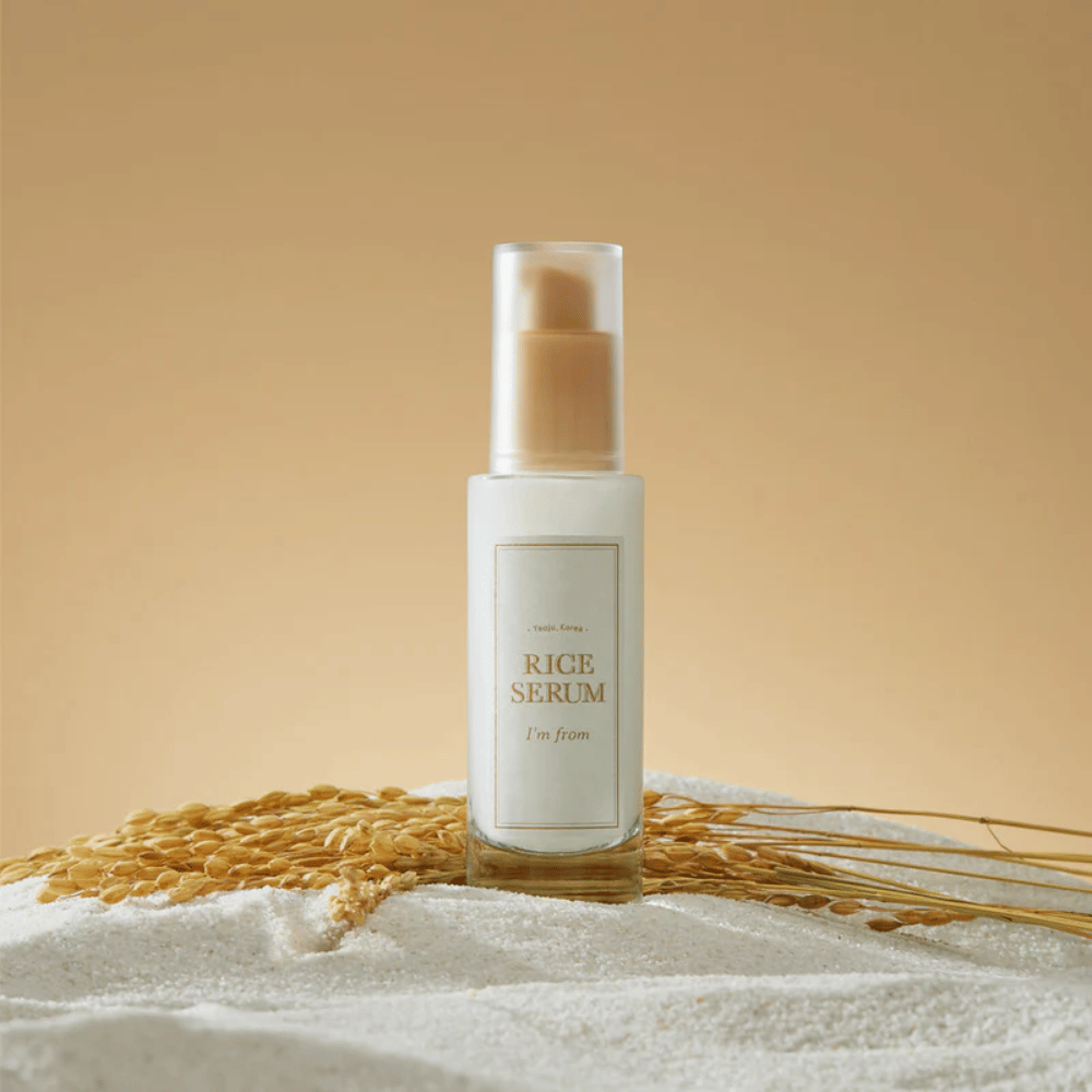 Flaske med "I'm from" Rice Serum, omgitt av riskorn og risaks mot en varm bakgrunn.