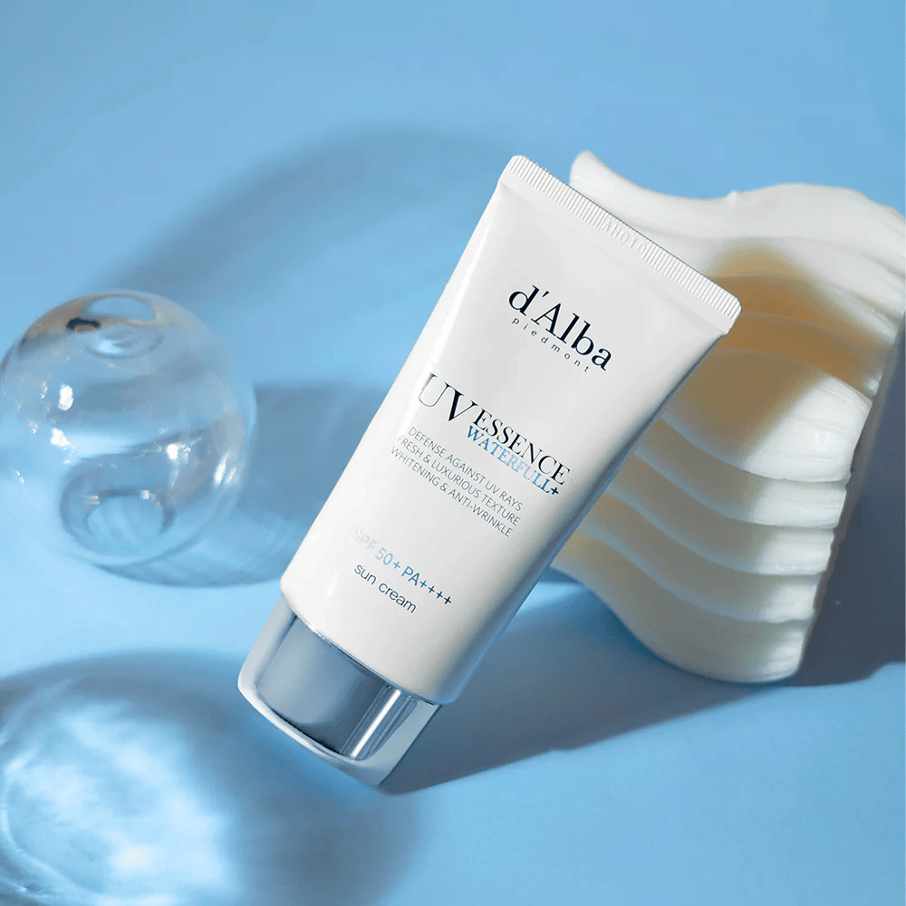 d'Alba UV Essence Waterfull+ Sun Cream med SPF 50+ PA++++ ligger mot en blå bakgrunn med lysrefleksjoner og dekorative elementer – fremhever solkremens friske og fuktighetsgivende egenskaper.