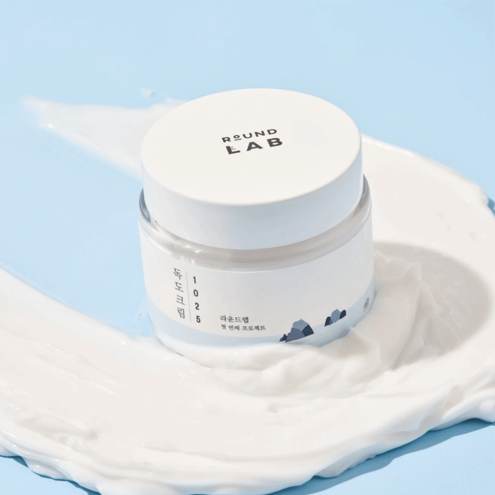 En krukke med Round Labs 1025 Dokdo Cream 80 ml som gir fuktighet, plassert på en levende blå bakgrunn.