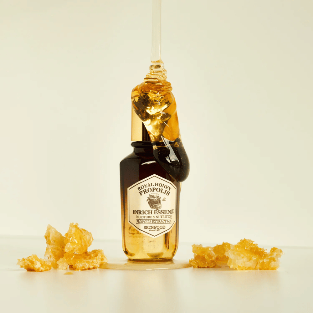 Bilde av en glassflaske med Royal Honey Propolis Enrich Essence fra Skinfood. En stor dråpe av gyllen honning renner ned fra flaskens gullfargede lokk. Rundt flasken ligger små biter av honningkake spredt på en lys overflate. Essensen markedsføres med fokus på fuktighet og næring, med 63% propolisekstrakt.
