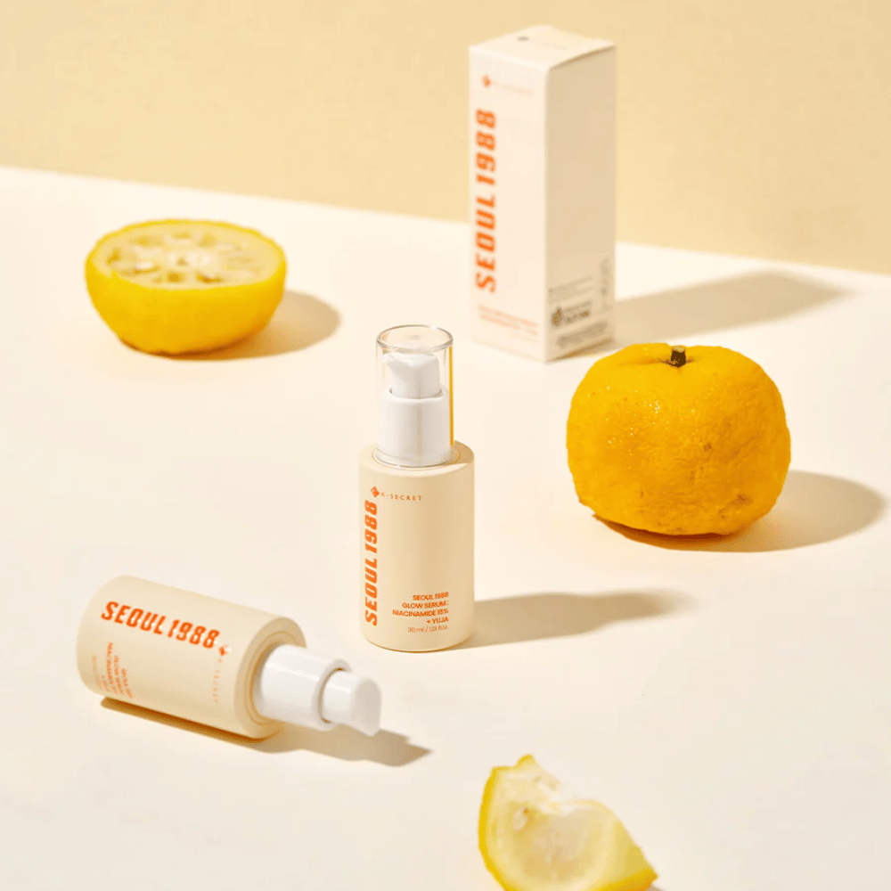 Flera flaskor och förpackningar av Seoul 1988 Glow Serum arrangerade med hela och halverade yuja-frukter i bakgrunden.