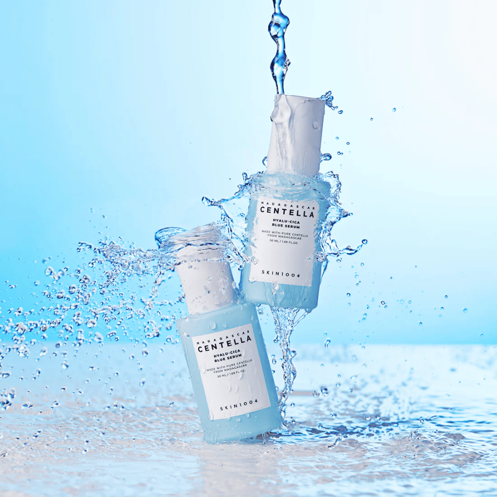 To flasker av SKIN1004 Madagascar Centella Hyalu-Cica Blue Serum treffes av en kaskade av vann mot en lys blå bakgrunn.