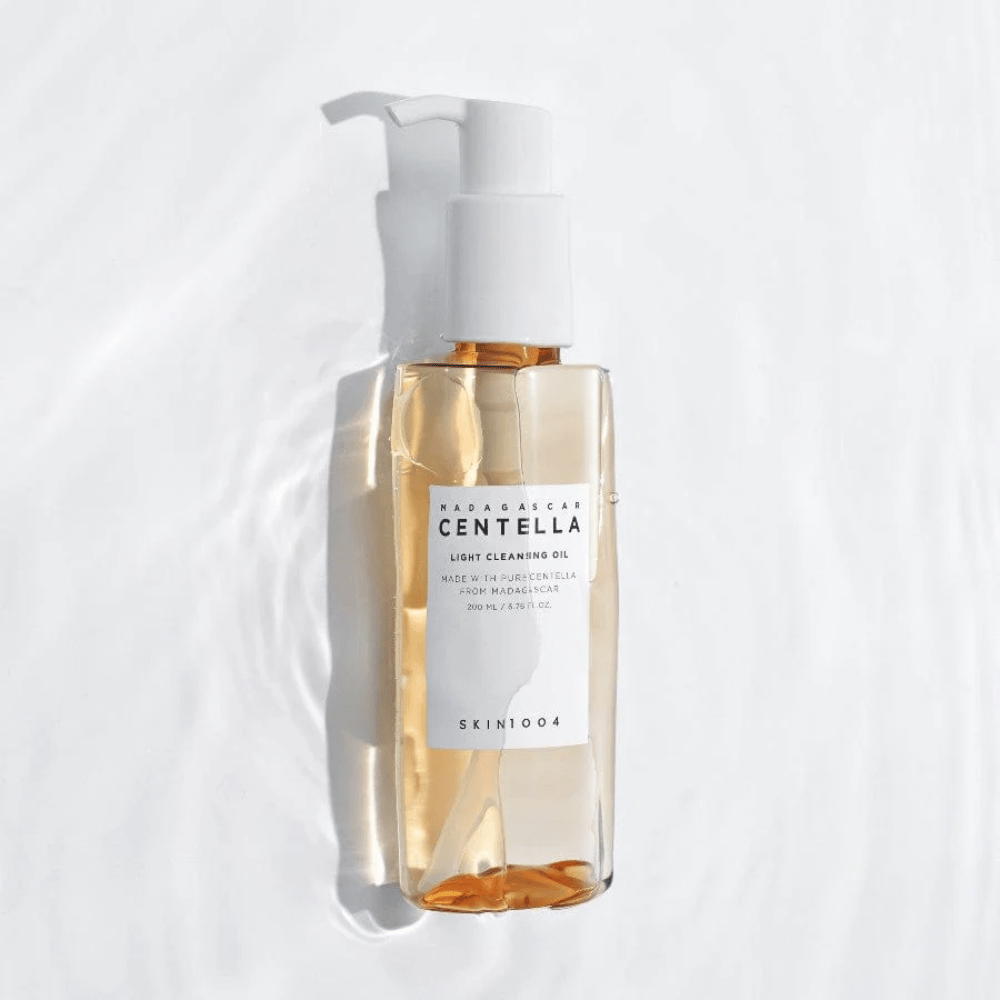 SKIN1004 Madagascar Centella Light Cleansing Oil på hvit bakgrunn med myke vannreflekser som fremhever den transparente flasken og gyldne innholdet.