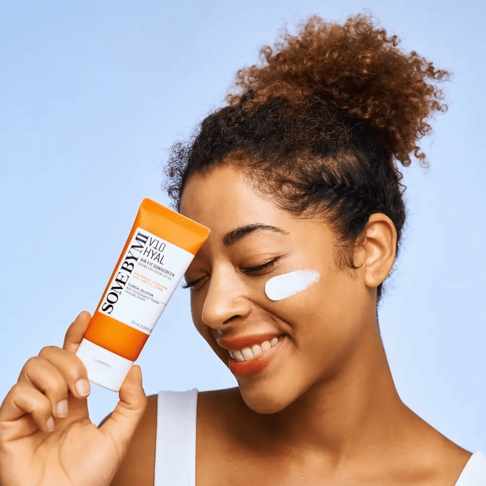 En kvinne holder en tube med SOME BY MI V10 Hyal Air Fit Sunscreen og har en hvit flekk av solkrem på kinnet. Hun smiler bredt og viser tydelig produktet, noe som fremhever dets bruk og fordeler for hudpleie og beskyttelse mot solen.