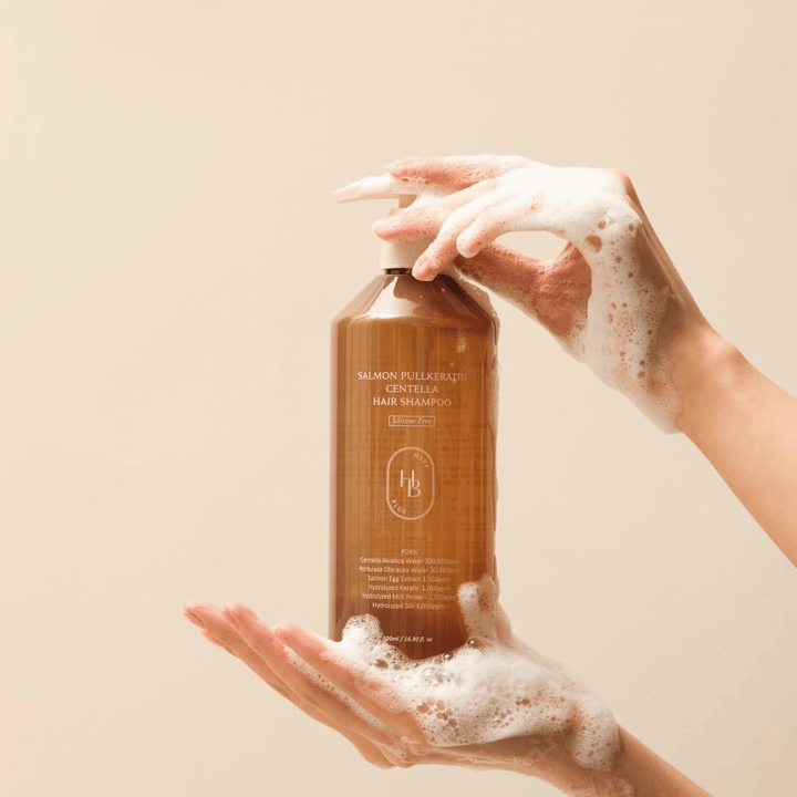 Heveblue Salmon Pullkeratin Centella Hair Shampoo i brun flaske omgitt av skum. Produktet holdes mellom to hender og fremhever en rensende og pleiende hårpleieopplevelse med rikelig skum.