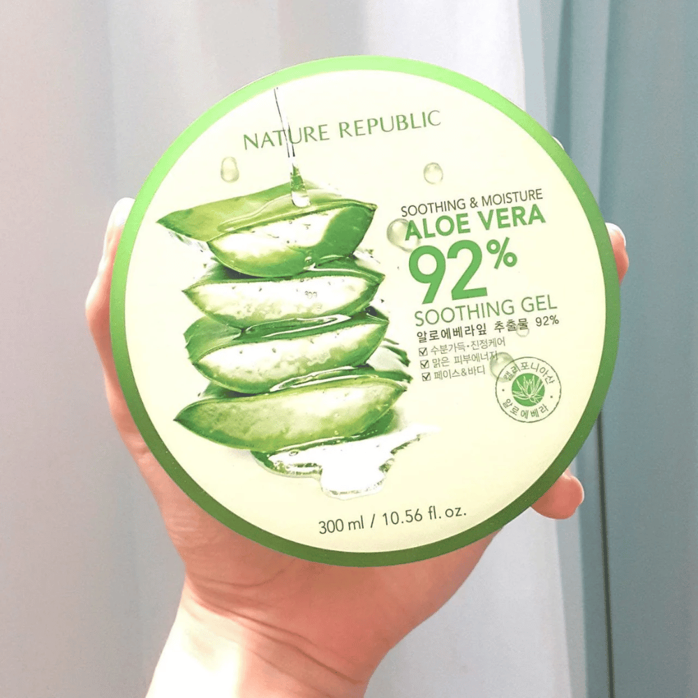En hånd holder opp en rund boks med Nature Republic Aloe Vera 92% Soothing Gel. Boksen er grønn med et bilde av aloe vera og tekst som beskriver dens beroligende og fuktighetsgivende egenskaper. Innholdet er 300 ml.