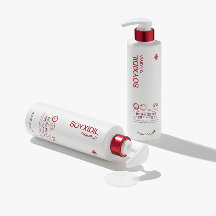 Två flaskor av Medicube Soyxidil Shampoo i vit och röd design, en stående och en liggande med schampo spillt bredvid.